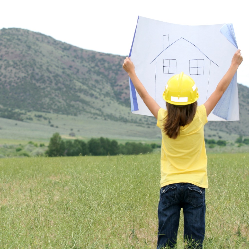 Child stand in an empty field with their back to the camera, wearing a hard hat and holding up a large drawing of a house